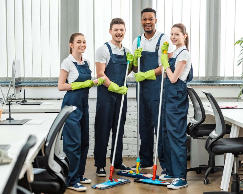 cheerful multicultural team of cleaners looking at camera while standing with mops in office cheerful multicultural team of cleaners looking at camera while standing with mops in office