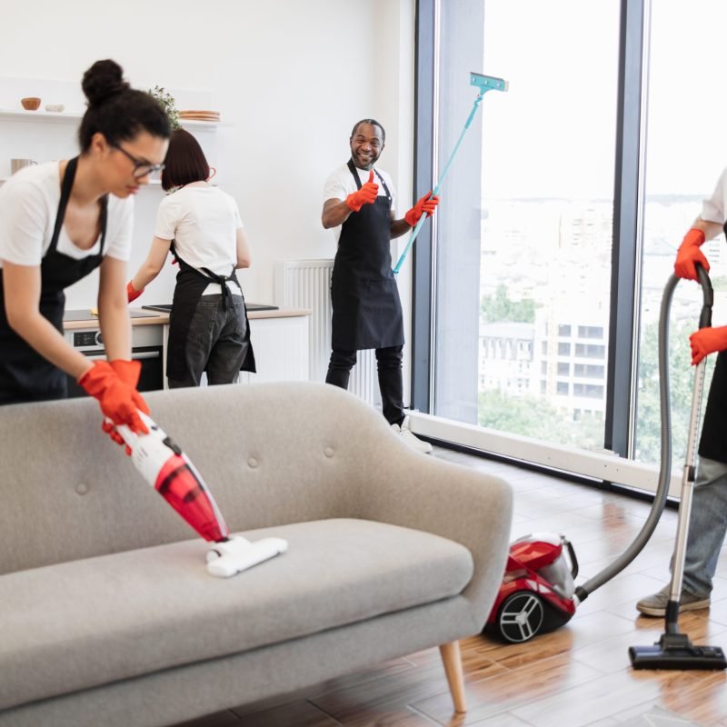 Focus on positive African American worker cleaning panoramic window. Focus on positive African American worker cleaning panoramic window glass with mop, looking at camera, showing thumb up in modern apartment near multiethnic co-workers.
