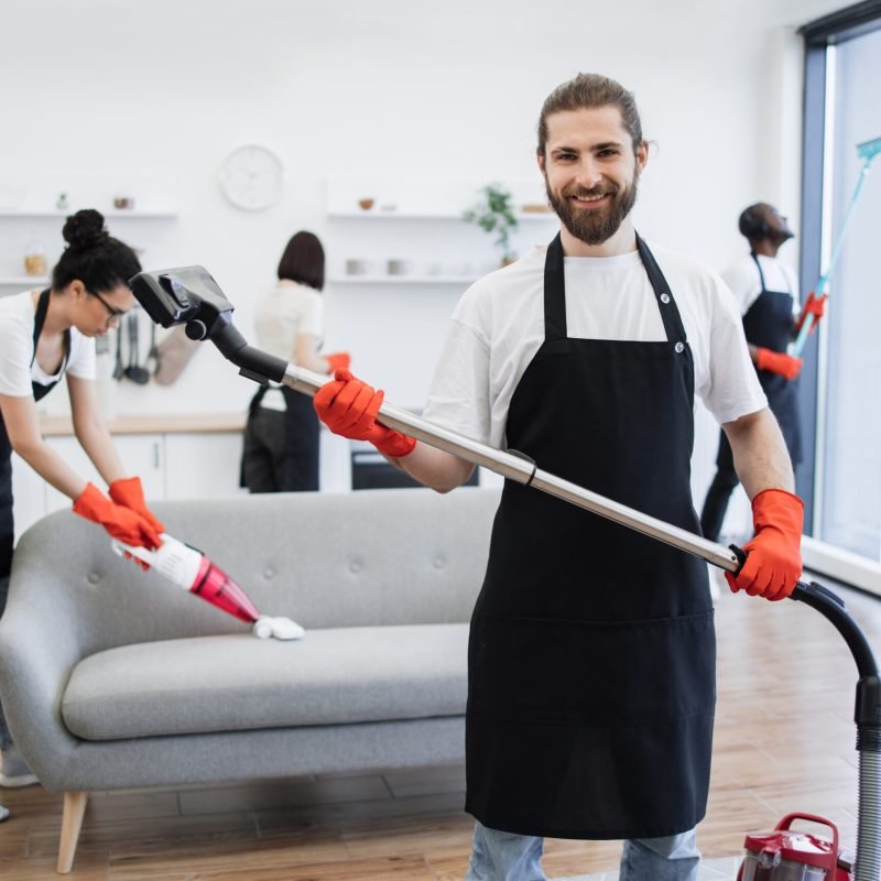 Portrait of bearded cleaner from cleaning service holding vacuum cleaner. Portrait of smiling bearded cleaner from cleaning service holding wireless vacuum cleaner in hands, looking at camera, against background of multicultural colleagues cleaning bright, modern kitchen.
